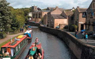 Narrowboat moves through a town with runners on one bank and other boats moored on the towpath