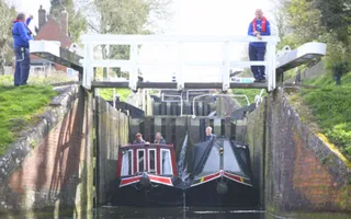 Two narrowboats at the bottom of a lock with the gates open and Canal & River Trust volunteers in life jackets stand on the bank.