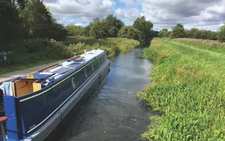 Narrowboat moves along a rural canal with vegetation sticking into the canal and a grassy towpath.