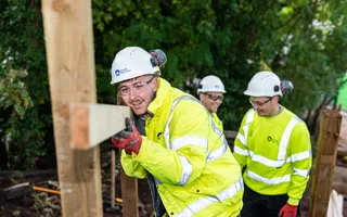 A teenage boy in a yellow hi-vis and white hard hat holds a horizontal plank of wood against vertical ones