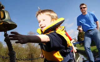 Child wearing a life jacket reaches for a bell on the back of a narrowboat