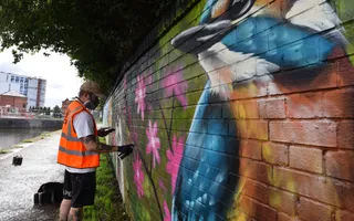 Man using spray cans to paint a mural of a kingfisher and wildflowers on a brick wall along a towpath