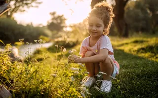 A young girl with dark curly hair in a ponytail and pink shorts and t-shirt smiles and crouches down next to the flowers on the towpath