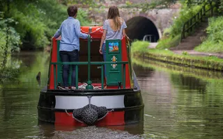 Two adults drive a narrowboat along the canal to a tunnel