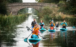 Group of people kneeling on paddleboards on a canal lined with soft verges and reeds.