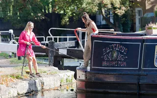 Woman pulling a narrowboat into the bank by a rope outside a lock while another person steers the boat