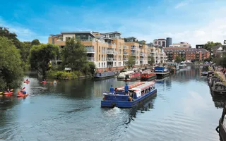 A boat travels away along a wide canal. To the left people in red canoes are paddling, and on the right a towpath is full of people walking. In the distance are blocks of flats and a blue sky