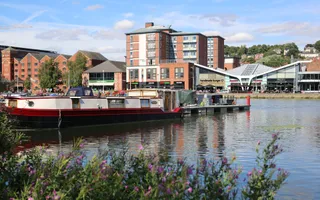 Picture of a canal boat on a river with buildings in the background and purple flowers in the foreground
