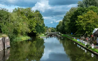 A calm canal with a series of lock gates in the distances is lined with large green trees, and reflects the blue sky on its surface.