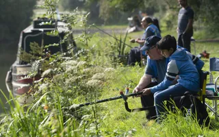 A young boy sits on a grassy towpath, holding a fishing rod with the help of an instructor.