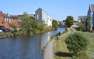 Wide shot of a canal on a sunny day with buildings on both sides and a couple of people on the towpath