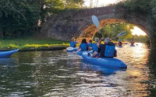 People in blue life jackets and blue canoes paddle away underneath a bridge, with the sun setting through it.