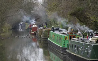 Boats line a tree-covered canal with smoke coming out of the chimneys.