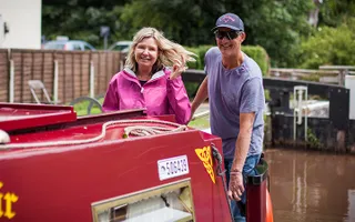 Man and woman stand on the back of a narrowboat, driving out of a lock