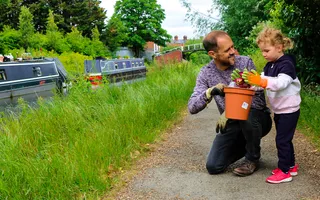 Man kneels on the towpath holding a plant pot for a child to fill