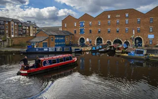 Red narrowboat approaches moored boats and industrial buildings