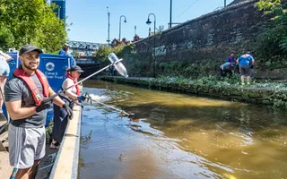 A man stands on a branded Canal & River Trust boat holding a litter picker with a plastic bottle in its pincer. A woman users a picker to collect more litter from the canal in the background.