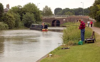 A man in a red and black checked shirt stands on a grassy towpath holding a rod out over the canal, whilst a boat chugs along in the distance towards a bridge.