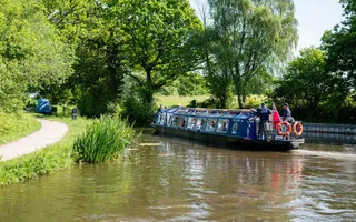 A blue narrowboat heads along a canal which is lined with a lush green towpath and trees