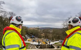 Two colleagues in high vis jackets and hard hats stand on a footbridge overlooking repair works on a lock in stormy weather.