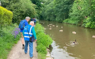 Two men in blue walk away from the camera along the towpath, looking down at geese on the canal to their right