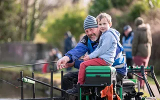 A young boy smiles at the camera as he learns to fish along the canal with an instructor