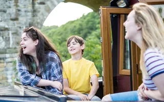 Group sit laughing in the cratch of a narrowboat as it passes a bridge