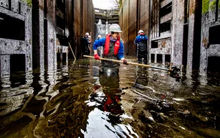 Volunteers in hard hats and life jackets stand in thigh-high water at the bottom of a lock removing debris from the canal bed
