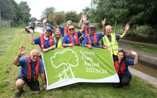Group of colleagues and volunteers smile with their arms raised, holding a sign saying "Green Flag Award 2023/24" by a lock.
