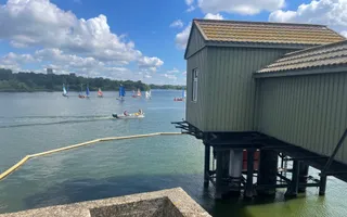 Light green building on stilts over water with sailing boats in the background on a sunny day
