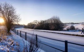 Man walks along snowy towpath next to an iced-over canal and snow-covered fields in the morning sun