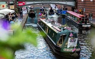 Narrowboat decorated with Union Jack bunting passes moored boats and pedestrians on a busy stretch of the canal, with bars and restaurants lining the towpath.