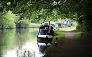 A line of boats moored on the towpath with people sitting on the grassy bank.
