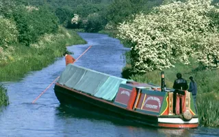 A person standing at the front of a narrowboat uses a pole to dislodge the boat which is grounded against the offside banm
